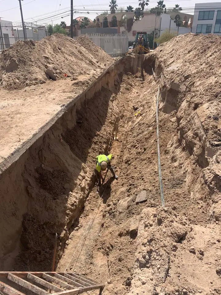 Zanja abierta a lo largo de la calle, con un trabajador afinando el fondo y la retroexcavadora al fondo