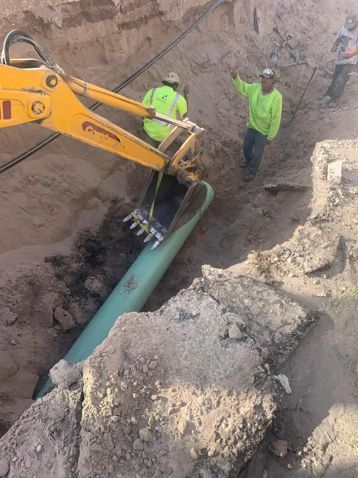 La retroexcavadora baja un tramo de tubería con eslinga mientras la cuadrilla guía la maniobra