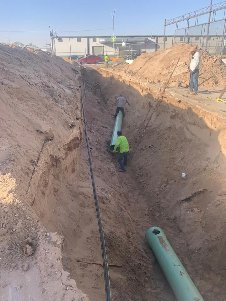 Trabajadores desplazando un tramo de tubería por el fondo de la zanja, con la planta industrial detrás